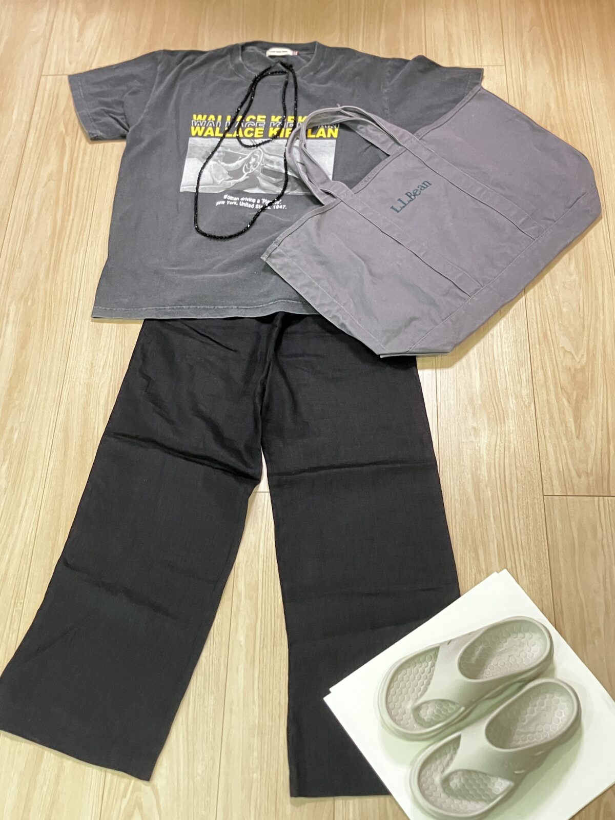 グレーのプリントTシャツ、黒のワイドパンツ、グレーのトートバッグ、白いサンダルを並べた移動日のコーデ